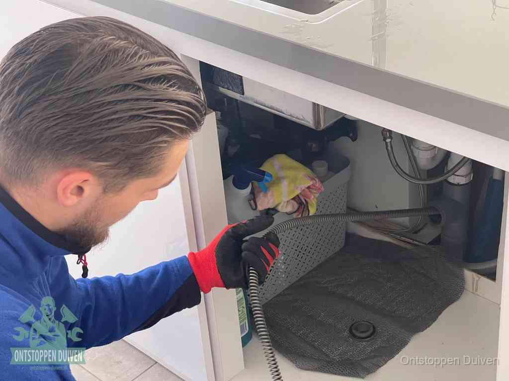 Een monteur voert een flexibele ontstoppingsveer in onder een keukenkastje om een verstopping voorbij de sifon te bereiken in Duiven