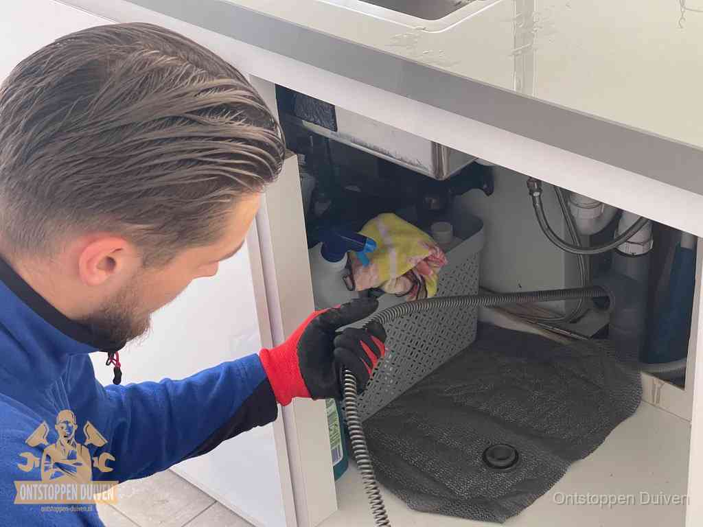Een monteur voert een flexibele ontstoppingsveer in onder het keukenkastje, langs de sifon, om de gootsteen te ontstoppen in Duiven Een monteur voert een flexibele ontstoppingsveer in onder het keukenkastje, langs de sifon, om de gootsteen te ontstoppen in Duiven
