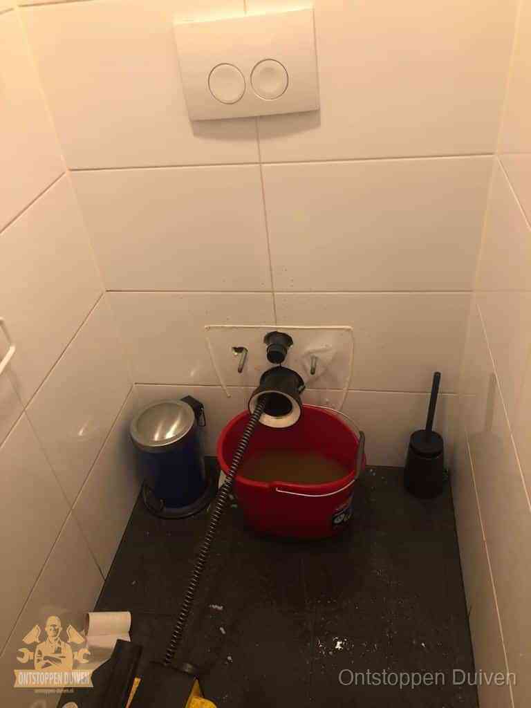 Een ontstopte wand-wc met verwijderde toiletpot waar een ontstoppingsveer in de wanduitgang verdwijnt, het achterhalen waar de verstopping zit in Duiven