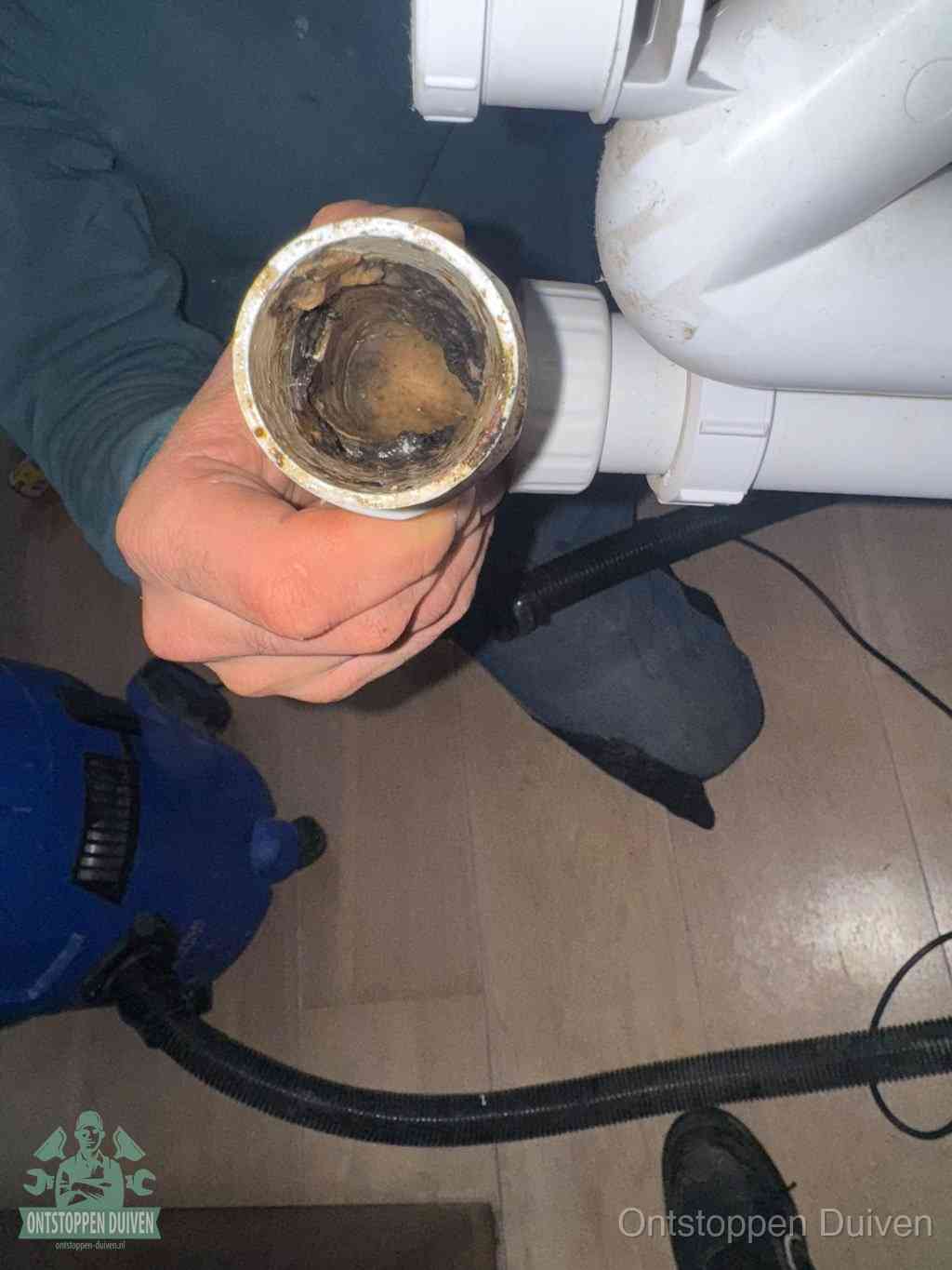 Een hand houdt een losgekoppeld PVC-afvoerbuisstuk vast dat van binnen volledig is dichtgeslibd met vet, vuil en aanslag, de meest voorkomende oorzaak van een verstopte afvoer in Duiven