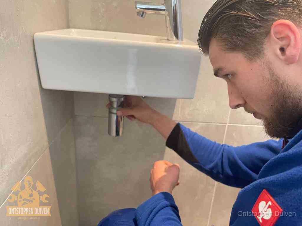 Een monteur draait met de hand de zwanenhalssifon onder een fonteinwasbak in de toiletruimte los om de bron van rioollucht in Duiven te vinden
