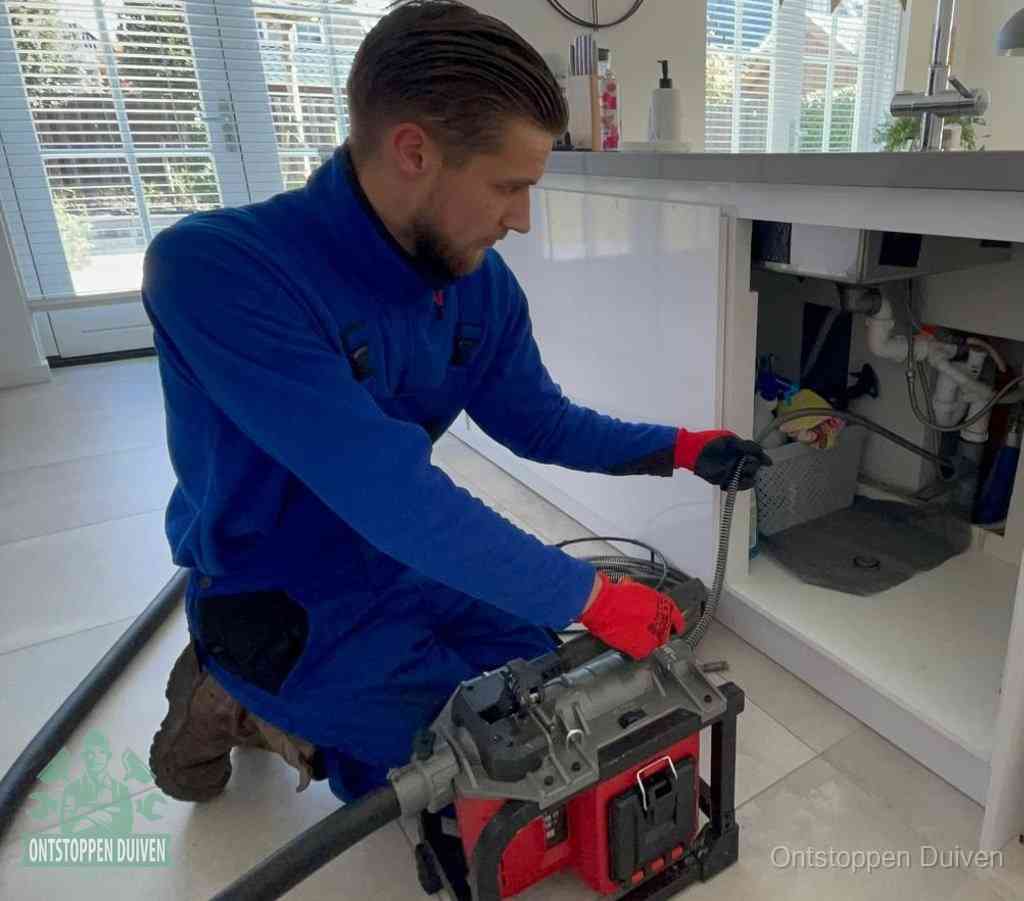 Een monteur met rode handschoenen knielt in een keuken en voert een elektrische ontstoppingsveer in tijdens het professioneel afvoer ontstoppen in Duiven